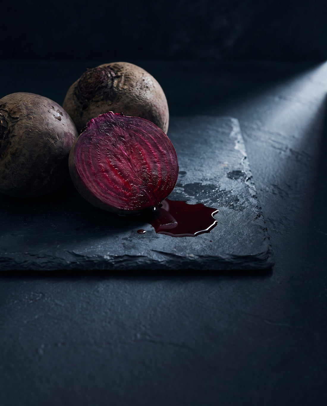 Whole and halved raw beetroot on dark slate, the cut face showing deep crimson rings