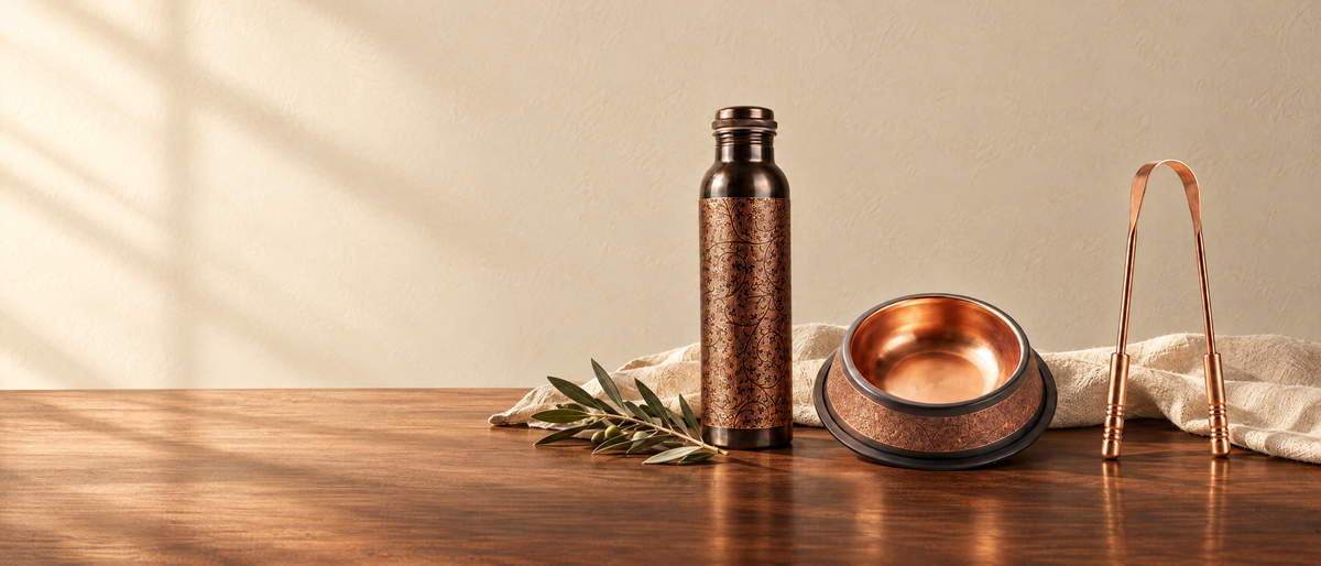 Etched copper water bottle, copper pet bowl and copper tongue cleaner on a walnut counter in morning light